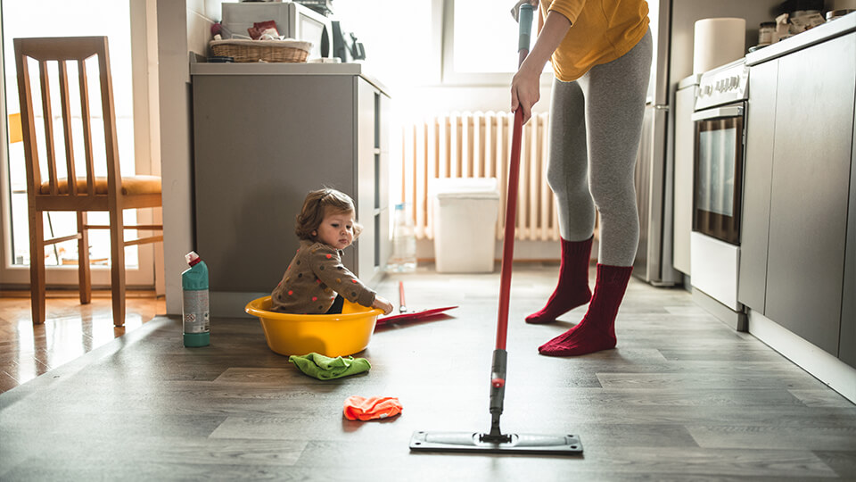 Mutter putzt die Wohnung während kleines Kind auf dem Boden sitzt.